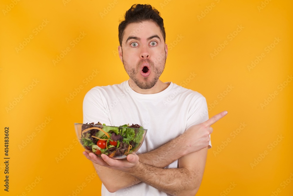 Confused young handsome Caucasian man holding a salad bowl against yellow wall chooses between two ways, points at both sides with crossed hands, feels doubt. Need your advice.
