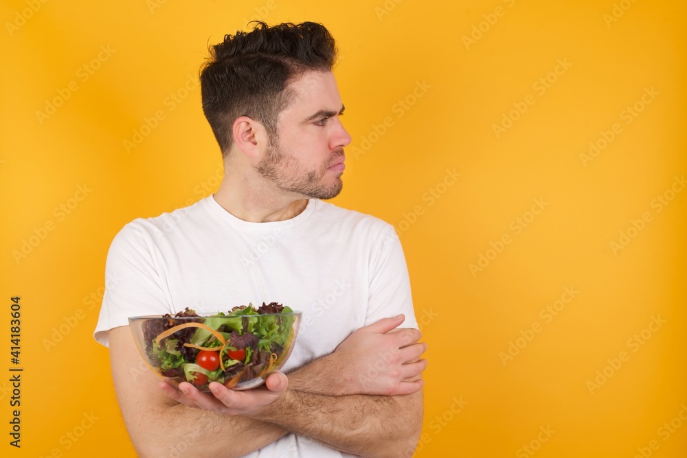 Image of upset young handsome Caucasian man holding a salad bowl against yellow wall with arms crossed. Looking with disappointed expression aside after listening to bad news.