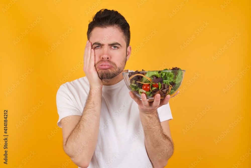 young handsome Caucasian man holding a salad bowl against yellow wall holding head in hands with unhappy expression watching sad movie about animals and trying not to cry. young handsome Caucasian man holding a salad bowl against yellow wall holding head in hands with unhappy expression watching sad movie about animals and trying not to cry.