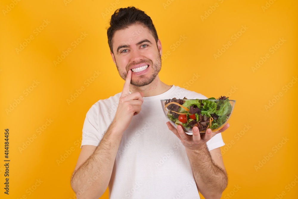 Carefree successful young handsome Caucasian man holding a salad bowl against yellow wall touching jawline gazing camera tilting head grinning white teeth delighted. Dental care concept. Carefree successful young handsome Caucasian man holding a salad bowl against yellow wall touching jawline gazing camera tilting head grinning white teeth delighted. Dental care concept.