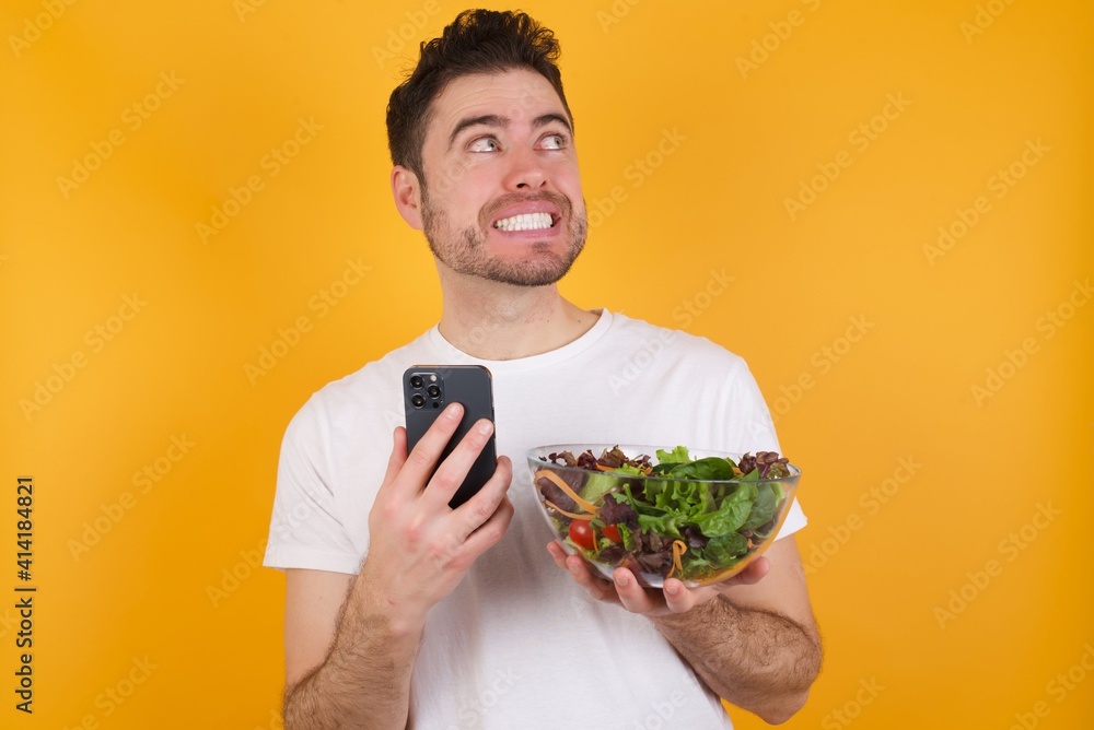 Image of a thinking dreaming young handsome Caucasian man holding a salad bowl against yellow wall using mobile phone and holding hand on face. Taking decisions and social media concept.