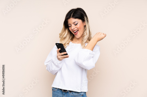 Teenager girl isolated on beige background with phone in victory position