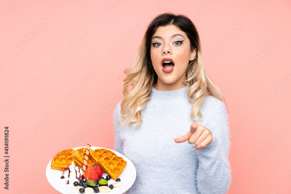 Teenager girl holding waffles on isolated pink background surprised and pointing front