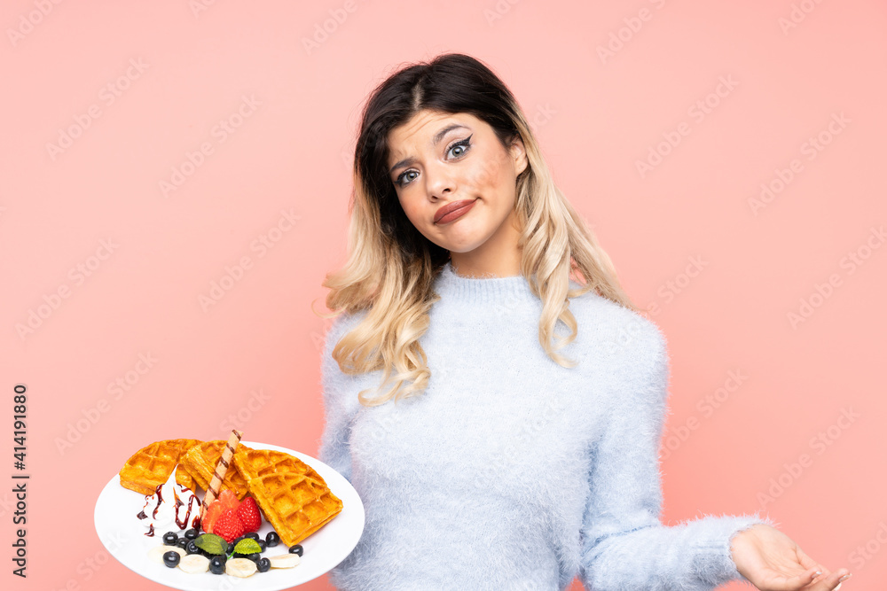 Teenager girl holding waffles on isolated pink background making doubts gesture while lifting the shoulders