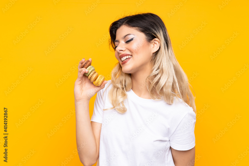 Teenager girl isolated on yellow background holding colorful French macarons and looking up while smiling Teenager girl isolated on yellow background holding colorful French macarons and looking up while smiling