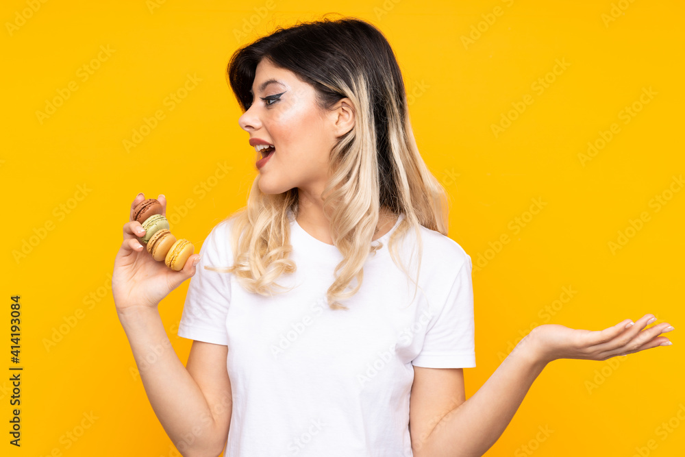 Teenager girl isolated on yellow background holding colorful French macarons and with surprise expression Teenager girl isolated on yellow background holding colorful French macarons and with surprise expression