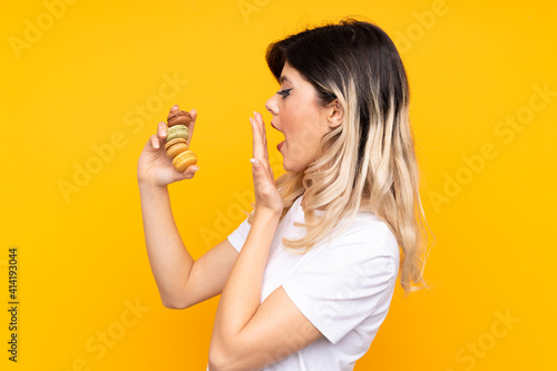 Teenager girl isolated on yellow background holding colorful French macarons and with surprise expression