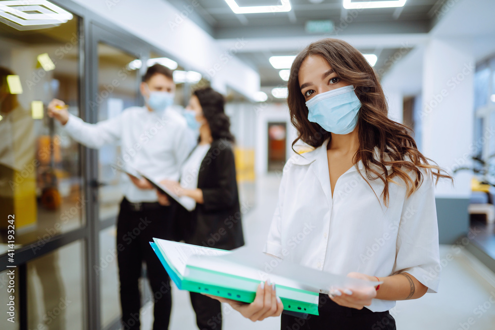 Obraz premium Portrait of young business woman in protective face mask an office building hallway. Office worker working during pandemic in quarantine city. COVID - 19.