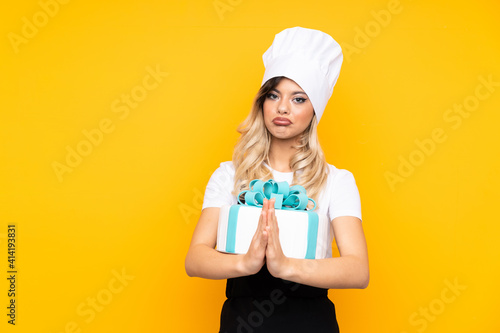 Teenager girl pastry holding a big cake isolated on yellow background pleading