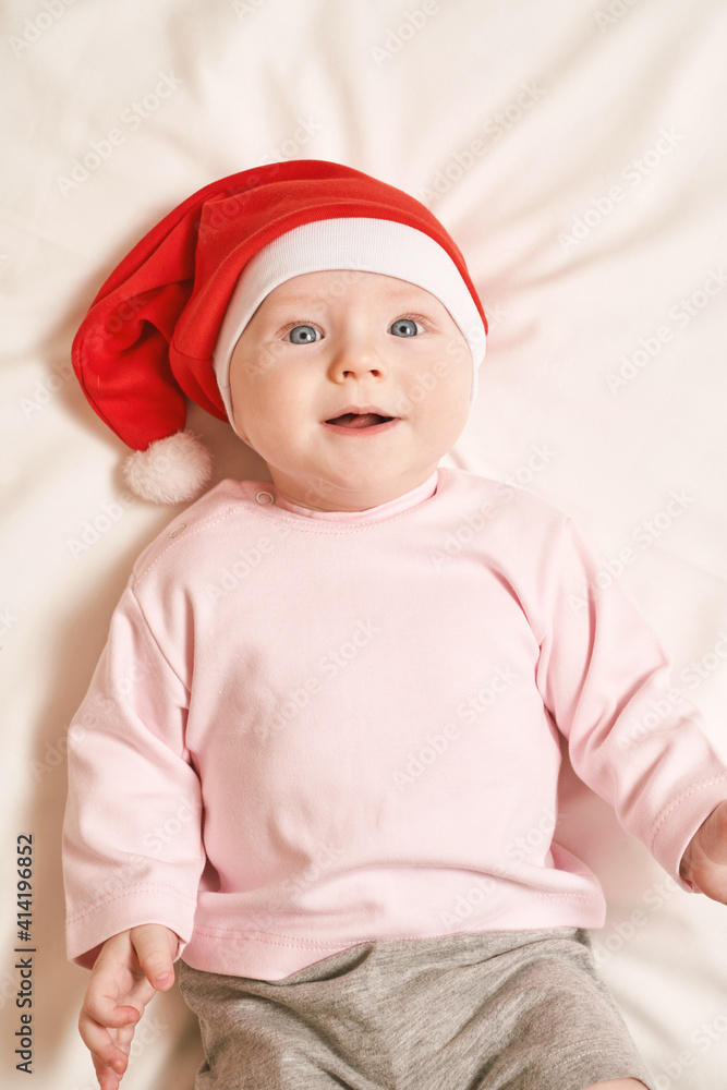Cute small boy lying at bed. Childhood christmas concept. Light background. Smiling child. Xmas red hat. Copyspace. Stay home. Mockup. Pink female clothes Cute small boy lying at bed. Childhood christmas concept. Light background. Smiling child. Xmas red hat. Copyspace. Stay home. Mockup. Pink female clothes
