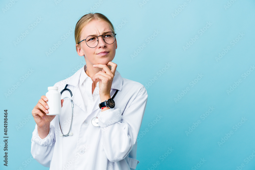 Young russian doctor woman holding pills bottle on blue looking sideways with doubtful and skeptical expression. Young russian doctor woman holding pills bottle on blue looking sideways with doubtful and skeptical expression.
