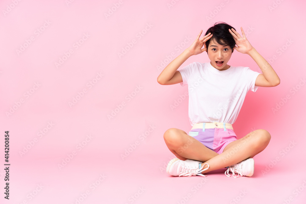 Young Vietnamese woman with short hair sitting on the floor over isolated pink background with surprise expression Young Vietnamese woman with short hair sitting on the floor over isolated pink background with surprise expression