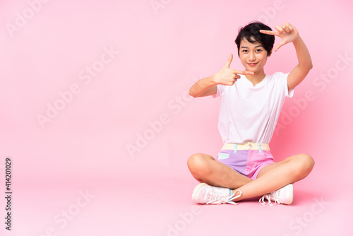 Young Vietnamese woman with short hair sitting on the floor over isolated pink background focusing face. Framing symbol