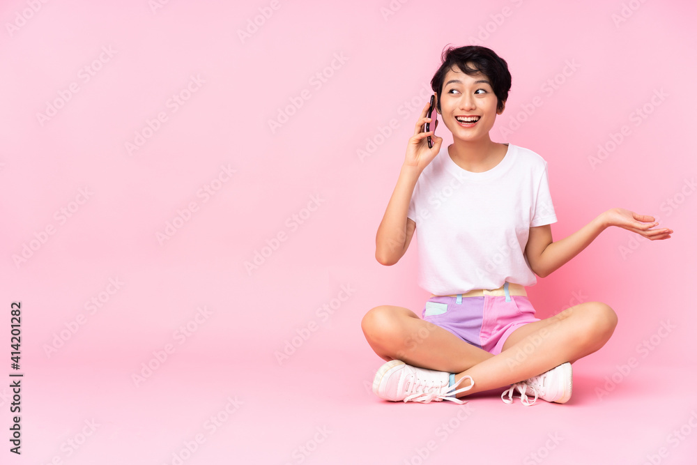Young Vietnamese woman with short hair sitting on the floor over isolated pink background keeping a conversation with the mobile phone with someone Young Vietnamese woman with short hair sitting on the floor over isolated pink background keeping a conversation with the mobile phone with someone