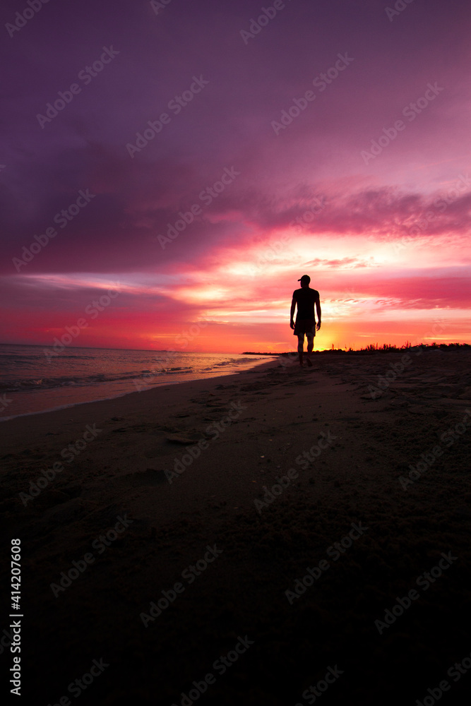 Persona a contraluz, caminando por la playa mientras el el amanecer de un hermoso cielo lo deja a oscuras. Persona a contraluz, caminando por la playa mientras el el amanecer de un hermoso cielo lo deja a oscuras.