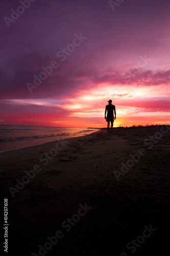 Persona a contraluz  caminando por la playa mientras el el amanecer de un hermoso cielo lo deja a oscuras.