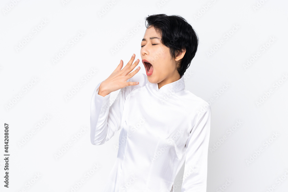Young Vietnamese woman with short hair wearing a traditional dress over isolated white background yawning and covering wide open mouth with hand Young Vietnamese woman with short hair wearing a traditional dress over isolated white background yawning and covering wide open mouth with hand