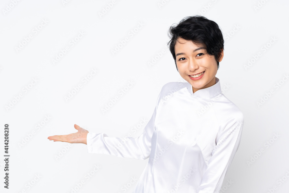 Young Vietnamese woman with short hair wearing a traditional dress over isolated white background extending hands to the side for inviting to come Young Vietnamese woman with short hair wearing a traditional dress over isolated white background extending hands to the side for inviting to come