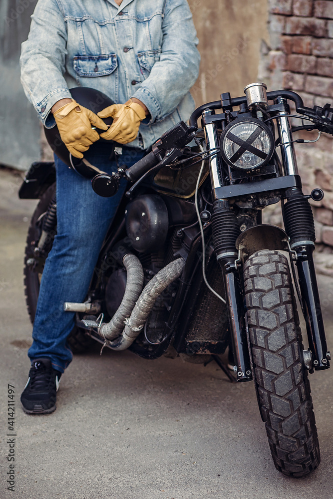 Brutal brave motorcycle driver in denim clothes and gloves holding a black helmet and sitting on a motorcycle. Motorcycles to order. The concept of masculinity Brutal brave motorcycle driver in denim clothes and gloves holding a black helmet and sitting on a motorcycle. Motorcycles to order. The concept of masculinity