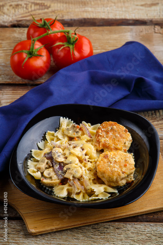 Pasta in a creamy sauce with mushrooms and chicken meatballs in a plate on a board on a blue napkin next to a tomato.