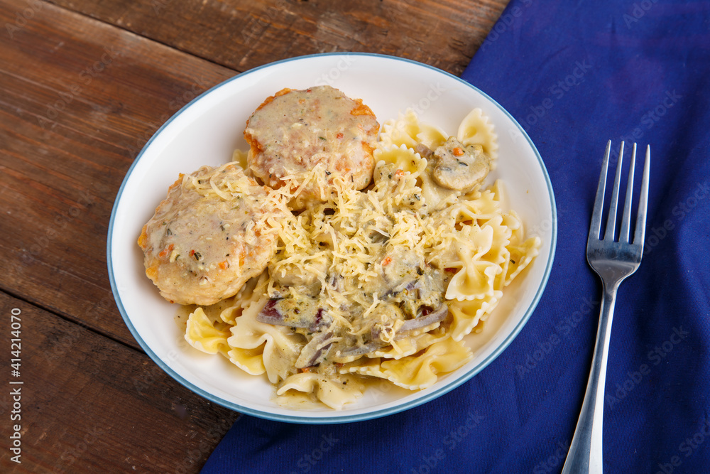 Pasta in a creamy sauce with mushrooms and chicken meatballs in a beige plate on a wooden table on a blue napkin next to a fork.