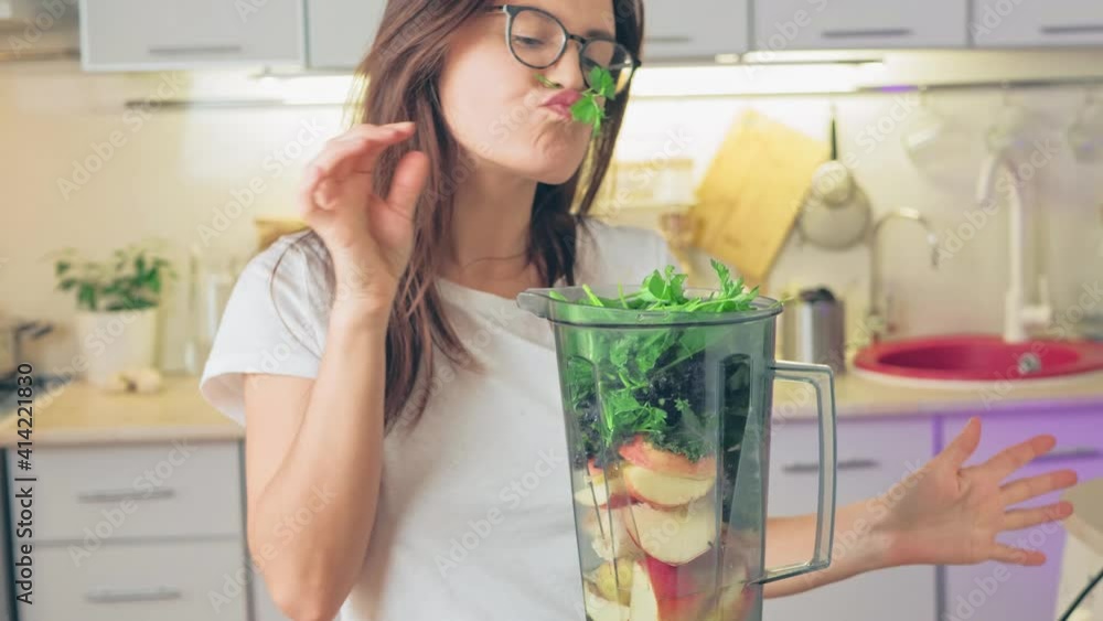 Funny young girl puts bunches of greens in a blender, plays with