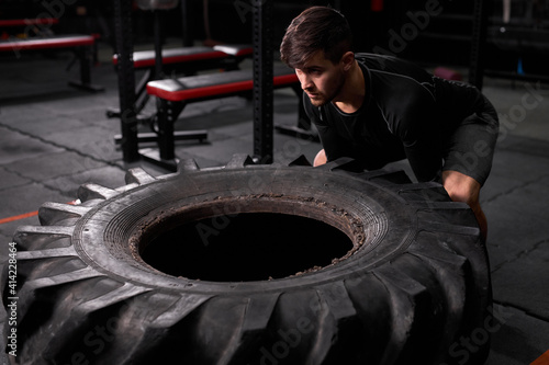 spotsman pushing drawn tire to build strength at modern fitness center. Male in sportswear is engaged in cross fit and workout. Sport, healthy lifestyle, bodybuilding, cross fit, workout concept photo