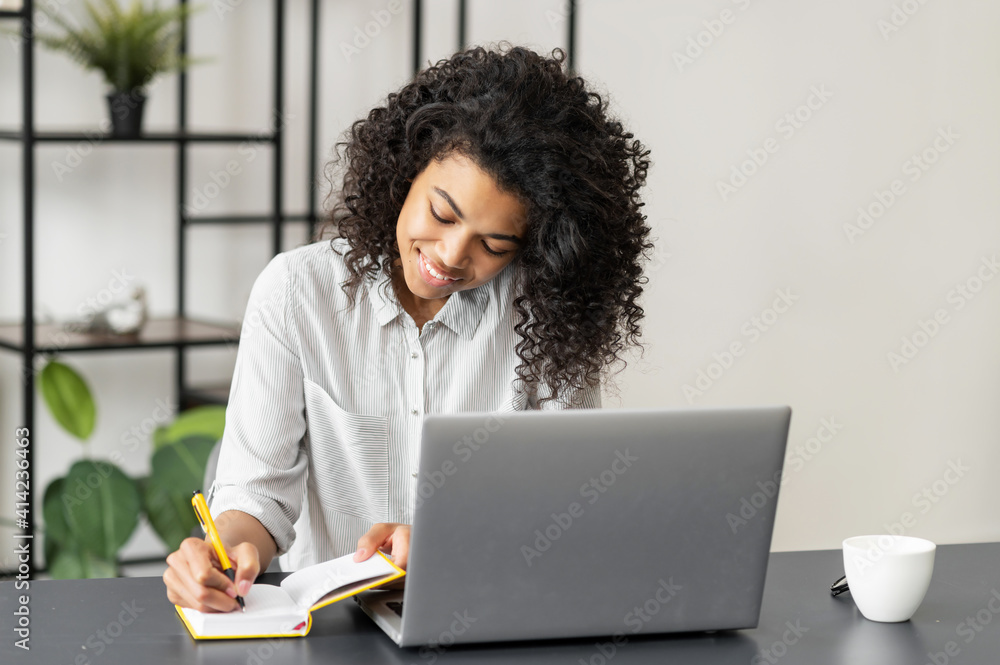 Positive African American woman student sitting at the desk at home in the modern office, using the laptop for distance video communication, studying, working, meeting online, and making notes Positive African American woman student sitting at the desk at home in the modern office, using the laptop for distance video communication, studying, working, meeting online, and making notes