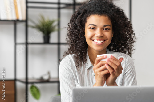 Smiling young cheerful African American woman with Afro hairstyle enjoying morning coffee while working on the laptop, sitting at the desk in the home office, happy businesswoman having coffee break