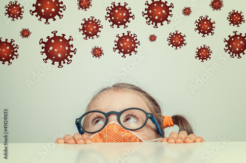 Prevention from Coronavirus concept: Beautiful cute little girl wearing protection mask while hiding under table and looking at Covid-19 virus illustrations above hear head.