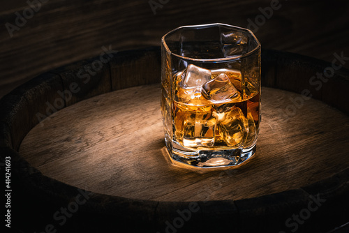 Glass of drink with a strong drink with ice on the background of an oak barrel with alcohol. Background with whiskey with ice in a glass close-up with copy space.