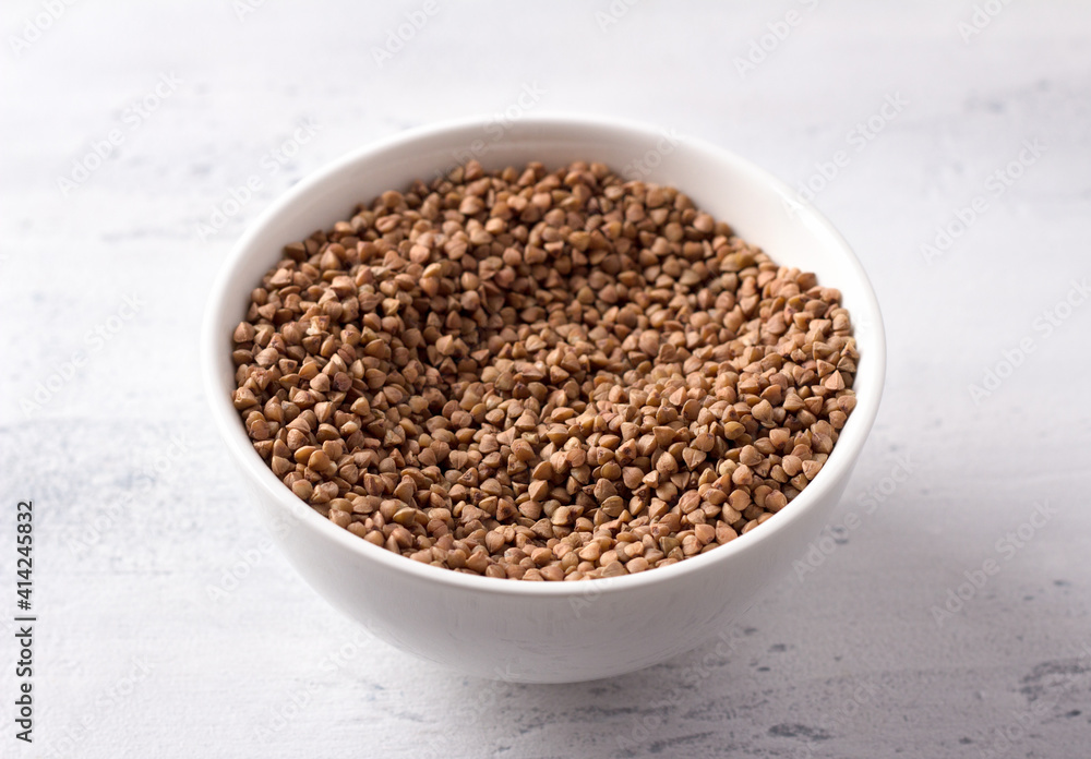 Raw buckwheat in a white ceramic bowl on a light gray textured background, horizontal, close up Raw buckwheat in a white ceramic bowl on a light gray textured background, horizontal, close up