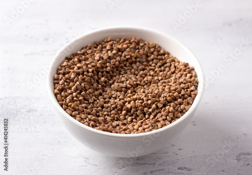 Raw buckwheat in a white ceramic bowl on a light gray textured background, horizontal, close up