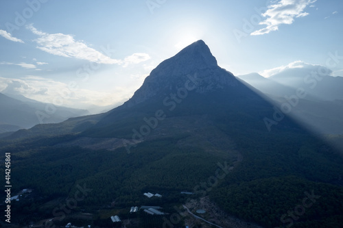 Sun shining through clouds and mountain peak. The view from the height of a green mountain valley with residential buildings surrounded by high mountains.