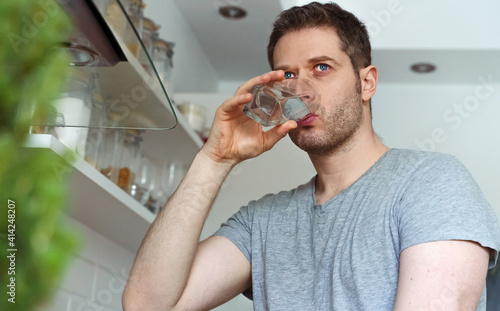Unshaven man drinks water from a glass in the kitchen.