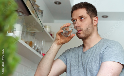 Unshaven man drinks water from a glass in the kitchen.