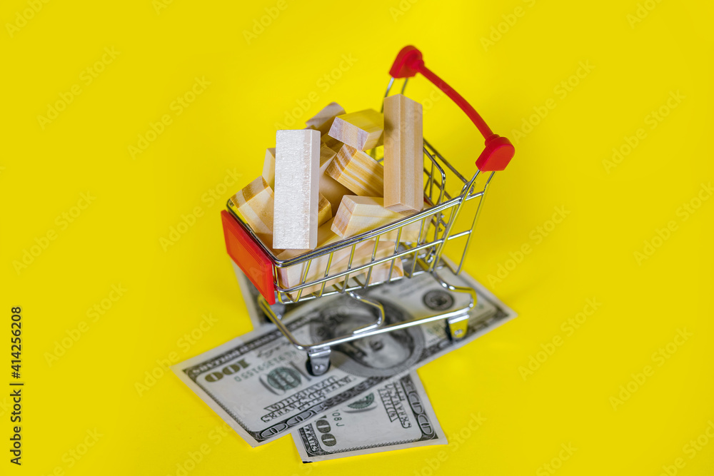 Trolley with wooden blocks and one hundred dollar bills on a yellow background. Real estate construction money spending concept top view