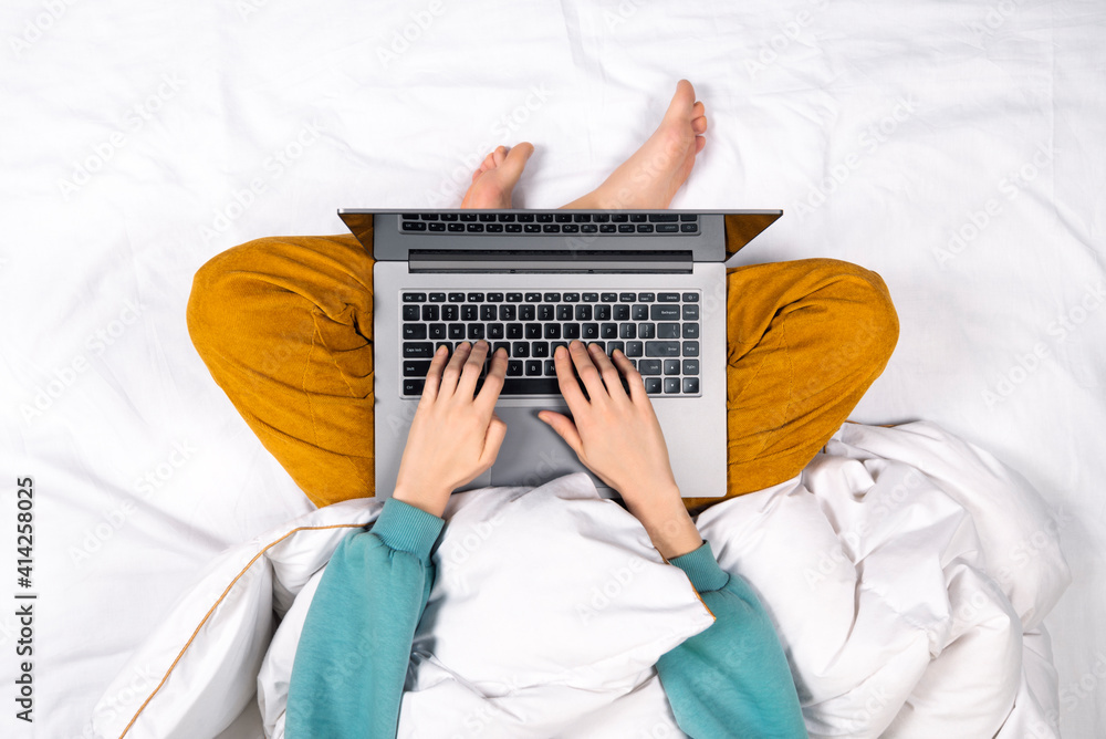 Fototapeta premium Top view on female hands working on modern laptop from bed at home. Home office or online education concept. Laptop lies on a woman's lap