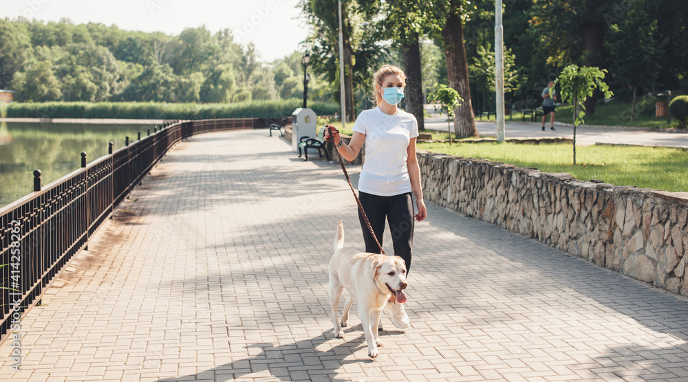 Blonde woman with medical mask is walking with her dog in park near a lake