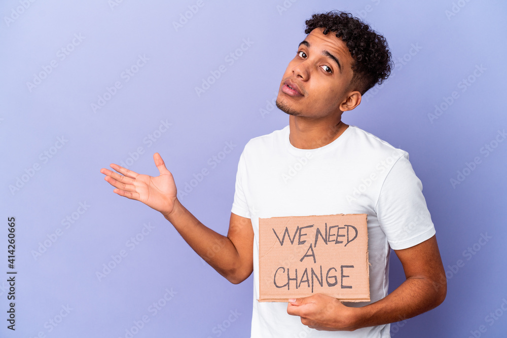 Young african american curly man isolated holding a we need a change cardboard showing a copy space on a palm and holding another hand on waist. Young african american curly man isolated holding a we need a change cardboard showing a copy space on a palm and holding another hand on waist.
