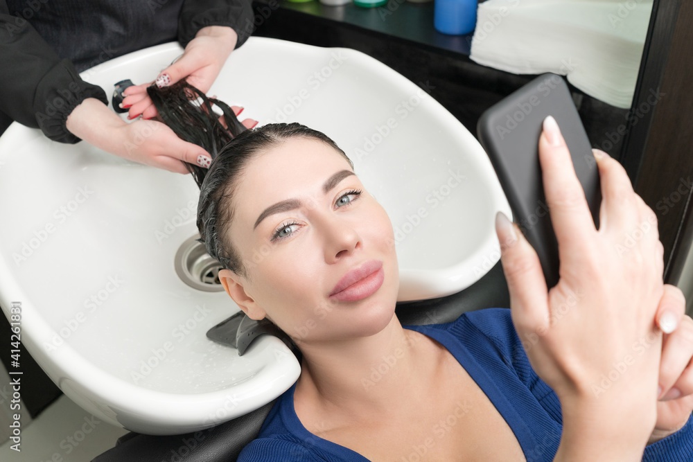 Fototapeta premium Brunette woman looks at smartphone while hairdresser is busy washing her hair at beauty salon. Hairstylist's hands wash long hair of customer with shampoo in sink for shampooing in beauty salon.