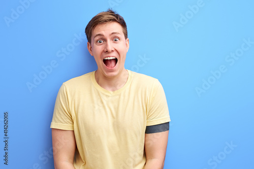 happy screaming male looking at camera isolated over blue background, handsome guy stand with wide opened mouth and eyes, feeling positive emotions photo