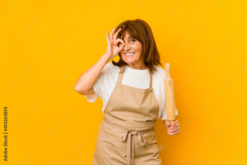 Middle age caucasian cook woman holding a roller isolated excited keeping ok gesture on eye. Middle age caucasian cook woman holding a roller isolated excited keeping ok gesture on eye.