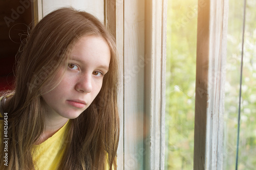beautiful girl with brown eyes, freckles and light brown hair looks at the camera. The girl at the window. Sad, unhappy teenager. Horizontal photo.