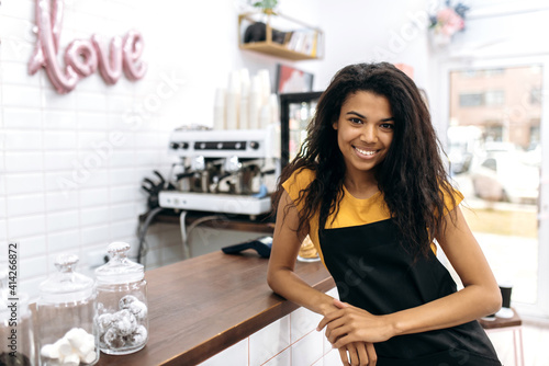 Photo of a friendly waitress wearing uniform, african american female barista, with curly hair in black apron standing near the bar counter, looks and smiles at the camera