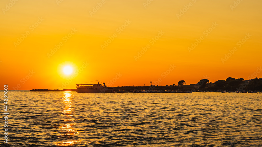 Amazing sunset with defined Sun and reflection on water. Silhouettes of boats and cruise ship moored at marina on Adriatic Sea, Zaton, Croatia