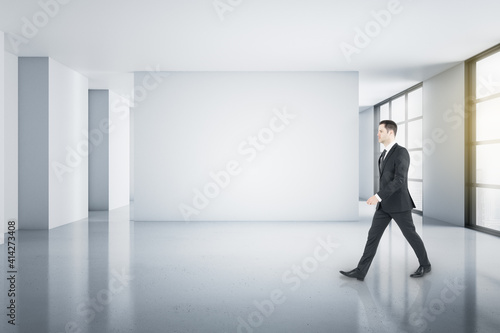 Young businessman walking in light spacious minimalistic room with blank light walls and windows to the floor. Mockup.