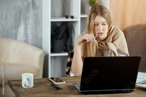 A focused student girl is preparing for a test exam doing homework with a ballpoint pen in her mouth at home sitting at a table with a laptop. The concept of self-education, distance learning.