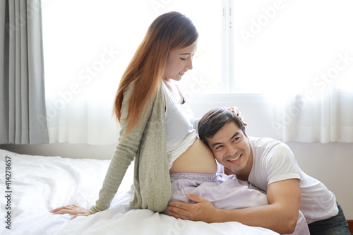 handsome caucasian husband listening his asian pregnant belly wife with happy smile face and say wow on bed in bedroom
