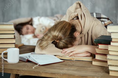 A student girl fell asleep over her books. Preparation for exams. A young girl fell asleep reading books at the table, in the background her friend is sleeping on the sofa covered with a blanket.
